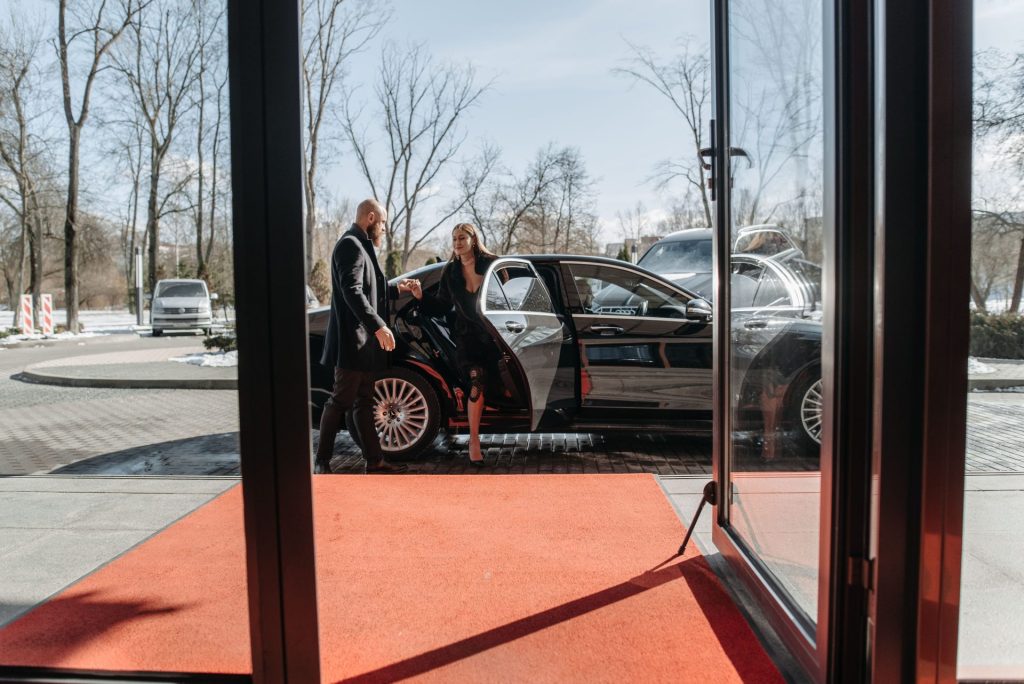 A couple stepping out of a luxury car onto a red carpet in an upscale outdoor setting.