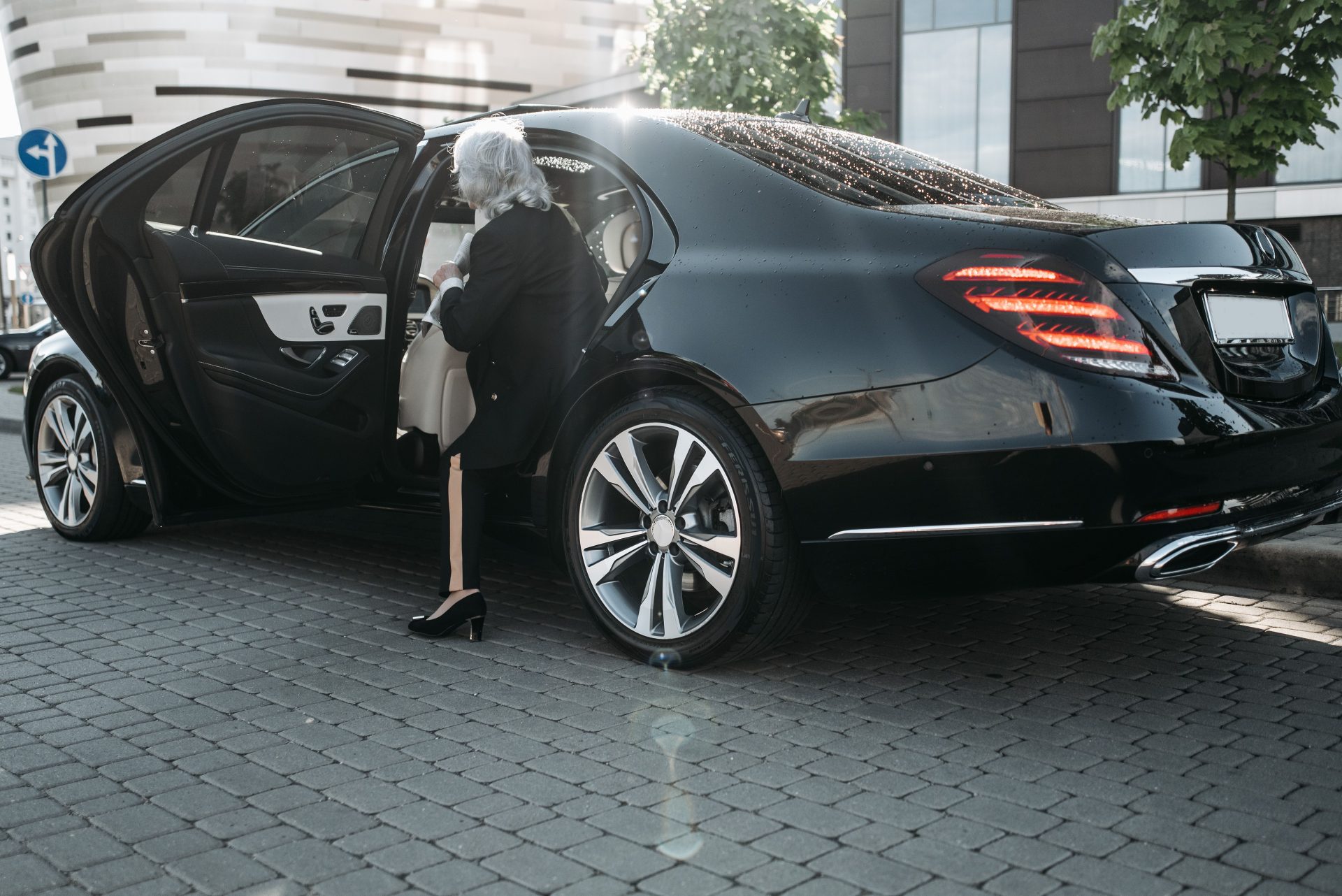 Senior businesswoman stepping out of a sleek black luxury vehicle in a cityscape.